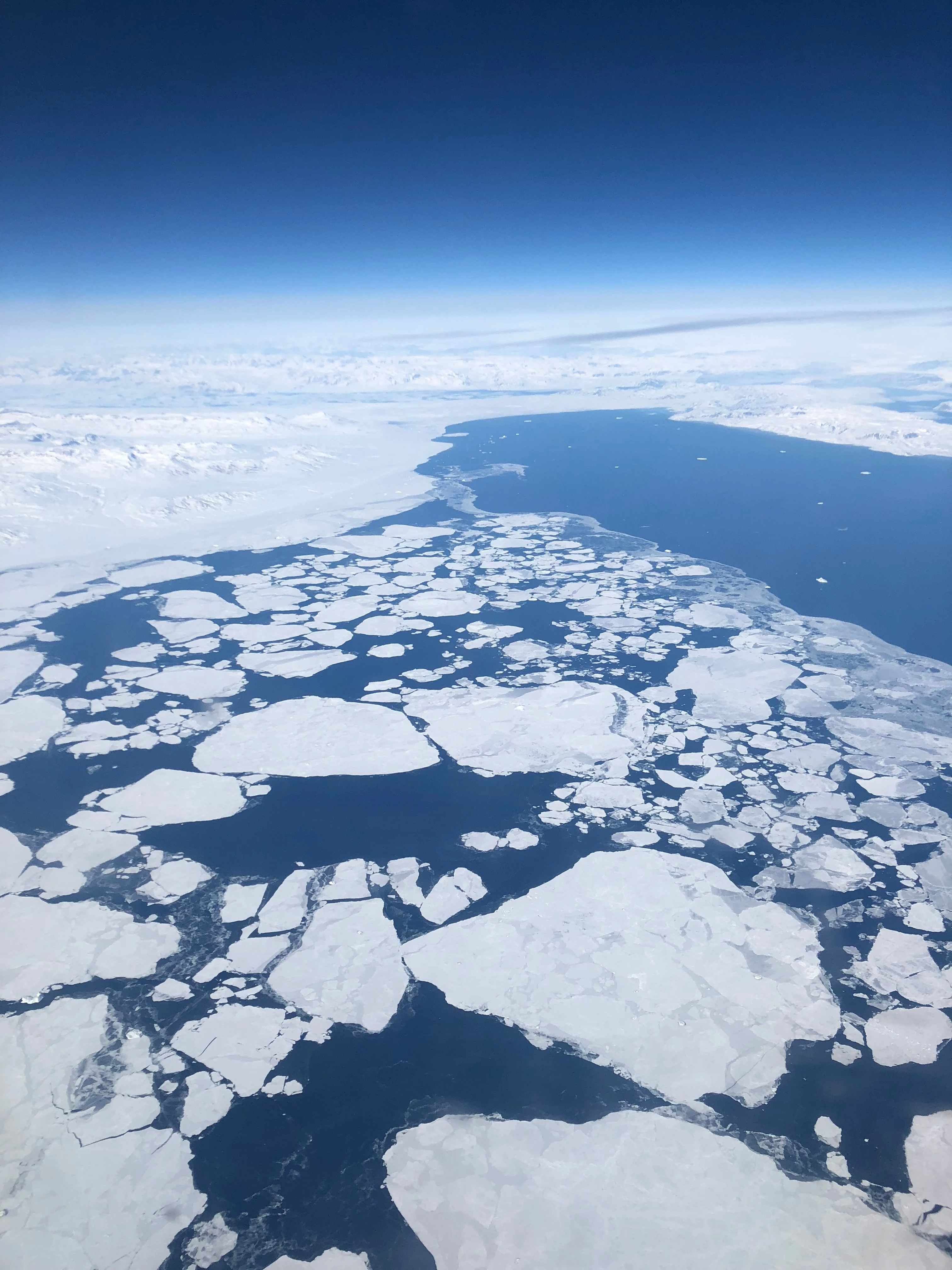 Broken white sea ice on dark blue water seen from high altitude, with a snow-covered coast beyond.